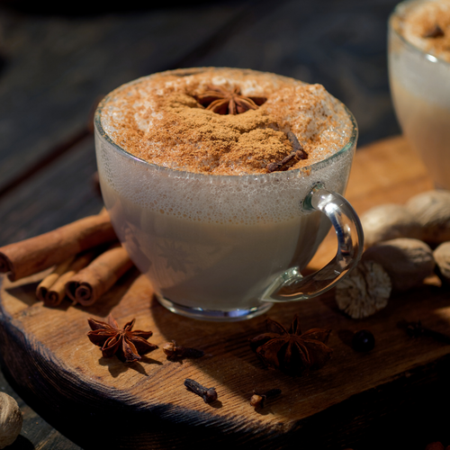 Vaso de cristal con chai latte espumoso, espolvoreado con canela y decorado con anís estrella y canela en rama sobre tabla.