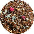 Mezcla de rooibos con trozos de fruta seca, pétalos rosas y hierbas verdes en fondo marrón.